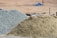 A large pile of sand and another pile of gravel are visible in an outdoor construction site. The background is a barren stretch of land with blurred objects and what appears to be construction debris.