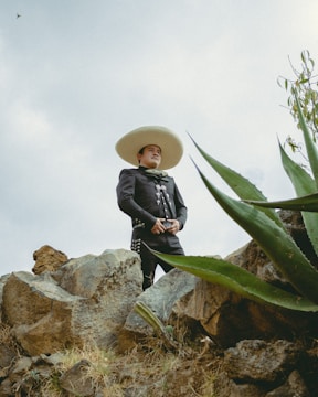A person wearing a traditional charro outfit with a large sombrero stands confidently on rocky terrain. The background features a cloudy sky, and large agave plants frame the foreground.