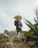 A person wearing a traditional charro outfit with a large sombrero stands confidently on rocky terrain. The background features a cloudy sky, and large agave plants frame the foreground.