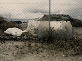 An abandoned area with a chain-link fence in the foreground. Behind the fence are large pieces of discarded white foam or plastic, surrounded by overgrown grass and shrubs. The sky is cloudy and the terrain looks barren, creating a sense of desolation or neglect.