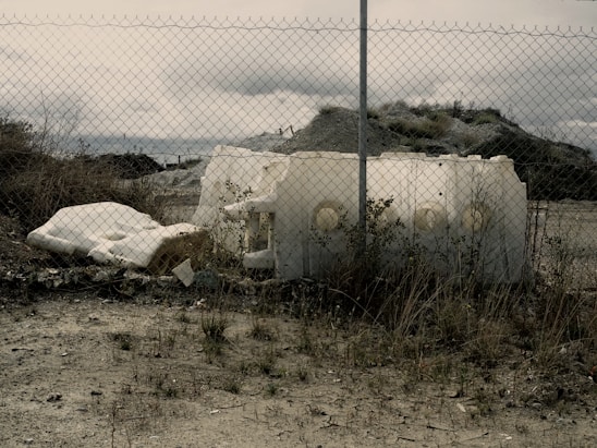 An abandoned area with a chain-link fence in the foreground. Behind the fence are large pieces of discarded white foam or plastic, surrounded by overgrown grass and shrubs. The sky is cloudy and the terrain looks barren, creating a sense of desolation or neglect.