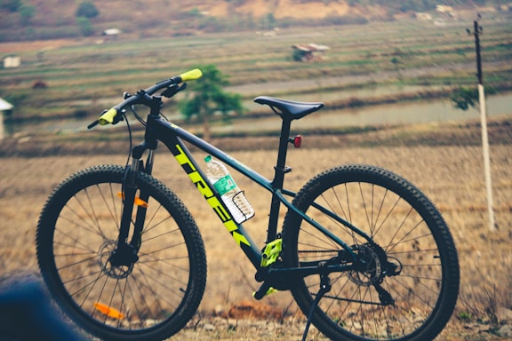 A mountain bike is positioned sideways with handlebars in the foreground, against a backdrop of terraced fields and distant hills. The bike frame is black with neon yellow and green accents, and there is a water bottle attached to the frame.