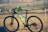 A mountain bike is positioned sideways with handlebars in the foreground, against a backdrop of terraced fields and distant hills. The bike frame is black with neon yellow and green accents, and there is a water bottle attached to the frame.