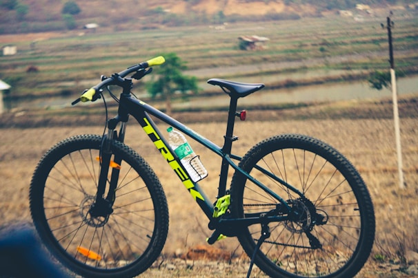 A mountain bike is positioned sideways with handlebars in the foreground, against a backdrop of terraced fields and distant hills. The bike frame is black with neon yellow and green accents, and there is a water bottle attached to the frame.