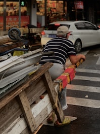 A person wearing a striped shirt and cap is sitting on a large wooden cart, loaded with various scrap materials. The cart is at the edge of a street lined with a pedestrian crossing. A white car is parked nearby, and in the background, there's a brightly lit storefront with signs and shelves displaying products.