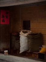 Several large bags filled with various items are stacked against a tiled wall. The bags are made of heavy fabric and are resting on top of plastic crates. There is a red poster with Chinese characters on the door to the left. A bucket is placed on the pavement in front of the bags.