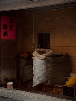 Several large bags filled with various items are stacked against a tiled wall. The bags are made of heavy fabric and are resting on top of plastic crates. There is a red poster with Chinese characters on the door to the left. A bucket is placed on the pavement in front of the bags.