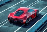 A sleek red sports car is parked on a racetrack with prominent aerodynamic design elements and a shiny finish. The car is viewed from the rear side, highlighting its detailed rear lights and large exhausts. Concrete barriers line the track, creating a controlled environment for high-speed testing.
