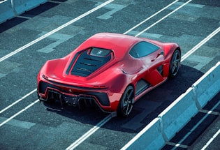 A sleek red sports car is parked on a racetrack with prominent aerodynamic design elements and a shiny finish. The car is viewed from the rear side, highlighting its detailed rear lights and large exhausts. Concrete barriers line the track, creating a controlled environment for high-speed testing.