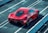 A sleek red sports car is parked on a racetrack with prominent aerodynamic design elements and a shiny finish. The car is viewed from the rear side, highlighting its detailed rear lights and large exhausts. Concrete barriers line the track, creating a controlled environment for high-speed testing.
