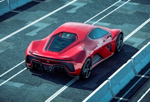 A sleek red sports car is parked on a racetrack with prominent aerodynamic design elements and a shiny finish. The car is viewed from the rear side, highlighting its detailed rear lights and large exhausts. Concrete barriers line the track, creating a controlled environment for high-speed testing.