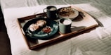 A breakfast tray is placed on a bed with sunlight streaming in. The tray holds two dark cups of frothy coffee, a black plate with pastries and a dollop of red jam, and a bowl of cereal with slices of fruit. The wooden tray contrasts with the crisp, white bedding.