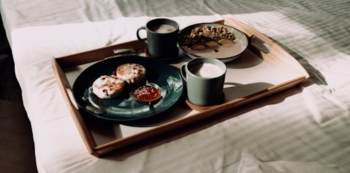 A breakfast tray is placed on a bed with sunlight streaming in. The tray holds two dark cups of frothy coffee, a black plate with pastries and a dollop of red jam, and a bowl of cereal with slices of fruit. The wooden tray contrasts with the crisp, white bedding.