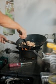 A person is cooking with a wok over a flame, stirring ingredients using a utensil. The kitchen setting includes nearby tools and ingredients with a subtle background decoration featuring a floral motif.