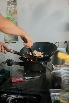 A person is cooking with a wok over a flame, stirring ingredients using a utensil. The kitchen setting includes nearby tools and ingredients with a subtle background decoration featuring a floral motif.