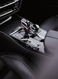 A sleek and modern car interior focused on the gear shift area, featuring a stylish gear lever with a red and blue accent. Surrounding the gear lever are various control buttons and a rotary knob, set within a glossy, black console. The seating appears to be made from high-quality leather with detailed stitching.