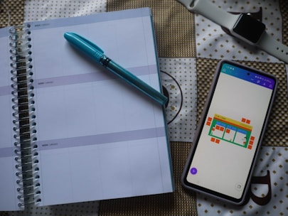 A spiral-bound planner is opened to a blank page with lines for daily entries. A blue pen is placed on top of the planner. Next to it, there is a smartphone with a colorful chart displayed on the screen. In the background, a smartwatch with a white strap is visible on a patterned surface.