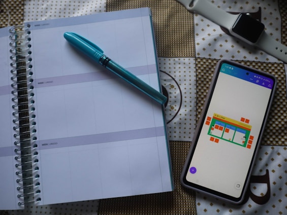 A spiral-bound planner is opened to a blank page with lines for daily entries. A blue pen is placed on top of the planner. Next to it, there is a smartphone with a colorful chart displayed on the screen. In the background, a smartwatch with a white strap is visible on a patterned surface.