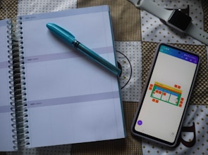 A spiral-bound planner is opened to a blank page with lines for daily entries. A blue pen is placed on top of the planner. Next to it, there is a smartphone with a colorful chart displayed on the screen. In the background, a smartwatch with a white strap is visible on a patterned surface.