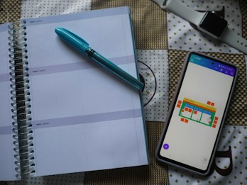 A spiral-bound planner is opened to a blank page with lines for daily entries. A blue pen is placed on top of the planner. Next to it, there is a smartphone with a colorful chart displayed on the screen. In the background, a smartwatch with a white strap is visible on a patterned surface.