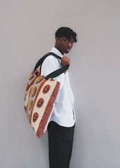 A person stands against a neutral background, looking slightly over their shoulder. They wear a white shirt and carry a large bag with African-inspired geometric patterns in warm tones.