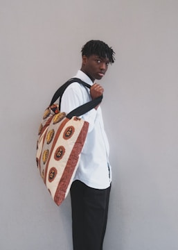 A person stands against a neutral background, looking slightly over their shoulder. They wear a white shirt and carry a large bag with African-inspired geometric patterns in warm tones.
