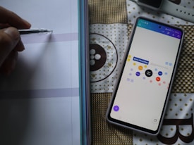 A hand is holding a pen poised to write on a notebook with lined paper. Beside it, a smartphone displays a digital mind map titled 'Story Mind Map', featuring various colored nodes. The background is a patterned fabric with geometric designs.