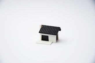 A small, minimalist model of a house with a black roof and a white base is centered on a plain white background. The house features a simple design with no additional details or surroundings.