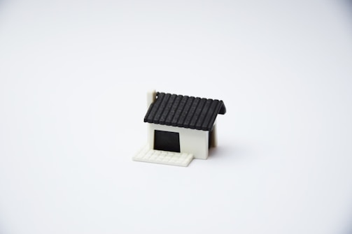 A small, minimalist model of a house with a black roof and a white base is centered on a plain white background. The house features a simple design with no additional details or surroundings.