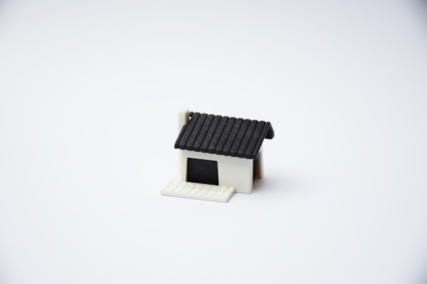 A small, minimalist model of a house with a black roof and a white base is centered on a plain white background. The house features a simple design with no additional details or surroundings.
