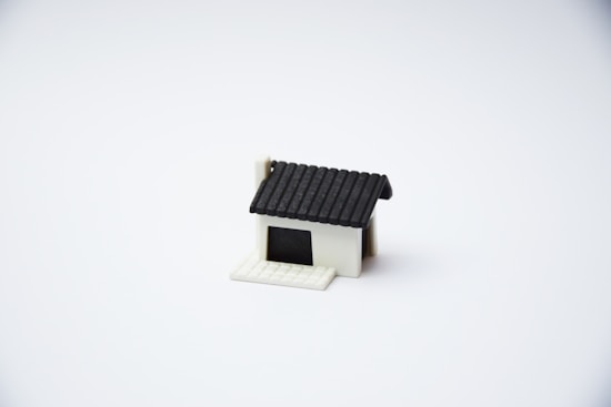 A small, minimalist model of a house with a black roof and a white base is centered on a plain white background. The house features a simple design with no additional details or surroundings.