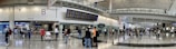 A bustling airport terminal with people walking and standing around, checking the departure and arrival boards. The spacious hall has high ceilings and polished floors, with various signs indicating gates and services. An information counter and foreign exchange booth are visible along one wall.