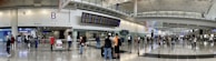 A bustling airport terminal with people walking and standing around, checking the departure and arrival boards. The spacious hall has high ceilings and polished floors, with various signs indicating gates and services. An information counter and foreign exchange booth are visible along one wall.