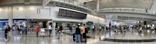 A bustling airport terminal with people walking and standing around, checking the departure and arrival boards. The spacious hall has high ceilings and polished floors, with various signs indicating gates and services. An information counter and foreign exchange booth are visible along one wall.