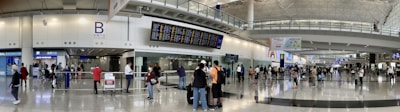 A bustling airport terminal with people walking and standing around, checking the departure and arrival boards. The spacious hall has high ceilings and polished floors, with various signs indicating gates and services. An information counter and foreign exchange booth are visible along one wall.