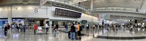 A bustling airport terminal with people walking and standing around, checking the departure and arrival boards. The spacious hall has high ceilings and polished floors, with various signs indicating gates and services. An information counter and foreign exchange booth are visible along one wall.