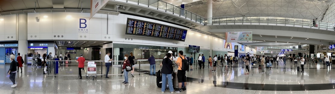 A bustling airport terminal with people walking and standing around, checking the departure and arrival boards. The spacious hall has high ceilings and polished floors, with various signs indicating gates and services. An information counter and foreign exchange booth are visible along one wall.
