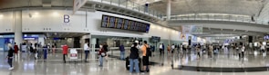 A bustling airport terminal with people walking and standing around, checking the departure and arrival boards. The spacious hall has high ceilings and polished floors, with various signs indicating gates and services. An information counter and foreign exchange booth are visible along one wall.