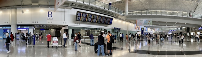 A bustling airport terminal with people walking and standing around, checking the departure and arrival boards. The spacious hall has high ceilings and polished floors, with various signs indicating gates and services. An information counter and foreign exchange booth are visible along one wall.