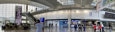 A busy airport terminal with people walking and sitting, featuring modern architecture with large windows allowing natural light. There are digital signage displays, an escalator, and a currency exchange booth visible.