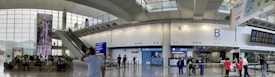 A busy airport terminal with people walking and sitting, featuring modern architecture with large windows allowing natural light. There are digital signage displays, an escalator, and a currency exchange booth visible.