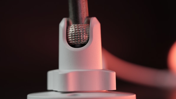 A close-up of a metallic component featuring a textured grid pattern, partially encased in a smooth white holder. The lighting casts a warm glow on the surface, highlighting details and creating a soft gradient in the background.