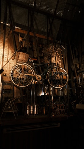 A vintage bicycle with a woven basket is suspended inside a rustic room. The backdrop features wooden elements and industrial metal beams, accompanied by various decorative objects and dried plants. Warm lighting creates a cozy and nostalgic atmosphere.