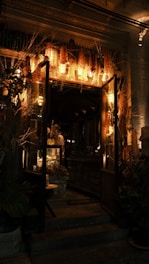 A warmly lit entrance of a vintage-style shop decorated with dried flowers and plants. The interior features wooden furniture and softly glowing lights, creating a cozy and inviting atmosphere.