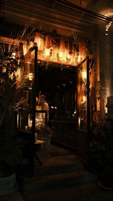 A warmly lit entrance of a vintage-style shop decorated with dried flowers and plants. The interior features wooden furniture and softly glowing lights, creating a cozy and inviting atmosphere.