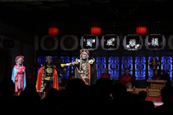 Performers in traditional Chinese opera costumes stand on stage under red lanterns. The background is adorned with blue and black decorative panels, and a group of musicians can be seen on the right. The atmosphere is dramatic and elaborate with a focus on vivid costumes and expressive masks.