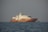 A large, elegant yacht with a wooden hull and white superstructure is floating on calm ocean waters. The weather appears clear with a slight haze near the horizon.