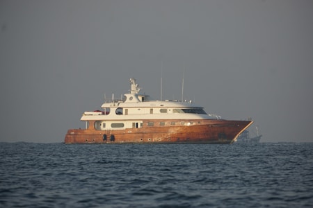 A large, elegant yacht with a wooden hull and white superstructure is floating on calm ocean waters. The weather appears clear with a slight haze near the horizon.