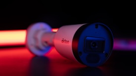 A surveillance camera is in focus, bathed in dramatic lighting with strong red and black tones. The brand name on the side is visible, and the camera's lens is slightly reflective. The background is dark, enhancing the illuminated effect of the camera.