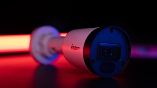 A surveillance camera is in focus, bathed in dramatic lighting with strong red and black tones. The brand name on the side is visible, and the camera's lens is slightly reflective. The background is dark, enhancing the illuminated effect of the camera.